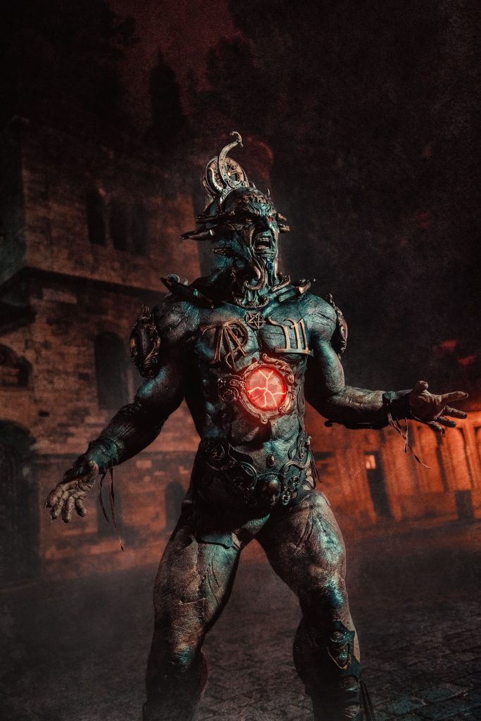 A vertical full-length shot of creature performer Jozef Rarach in the Golem prosthetic suit, standing with arms outstretched and a glowing red chest core at night outside the historic Jewish Ceremonial Hall next to the Klausen Synagogue in Prague.