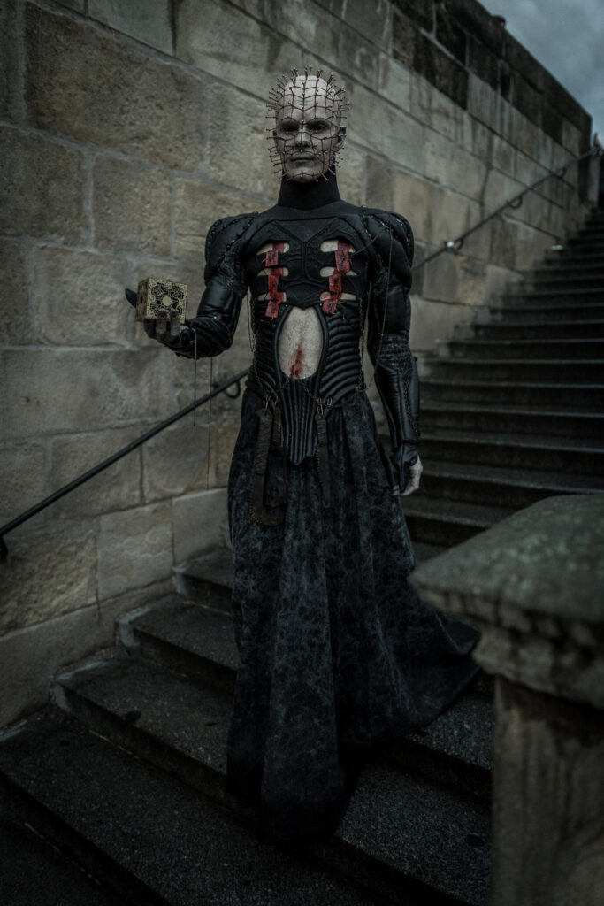A character with white prosthetic skin and metal pins covering his head in a grid pattern, standing on stone stairs and holding an ornate golden puzzle box.