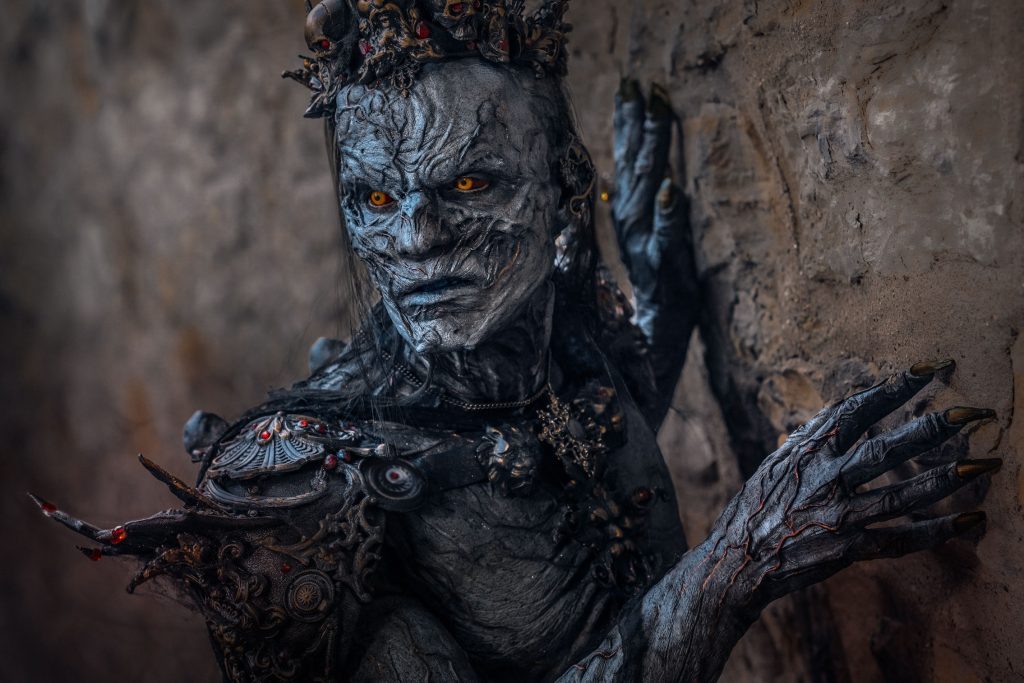 Close-up portrait of actor Jozef Rarach in full-body grey creature prosthetic makeup with glowing orange eyes and a crown, posing with clawed hands against an old stone wall.